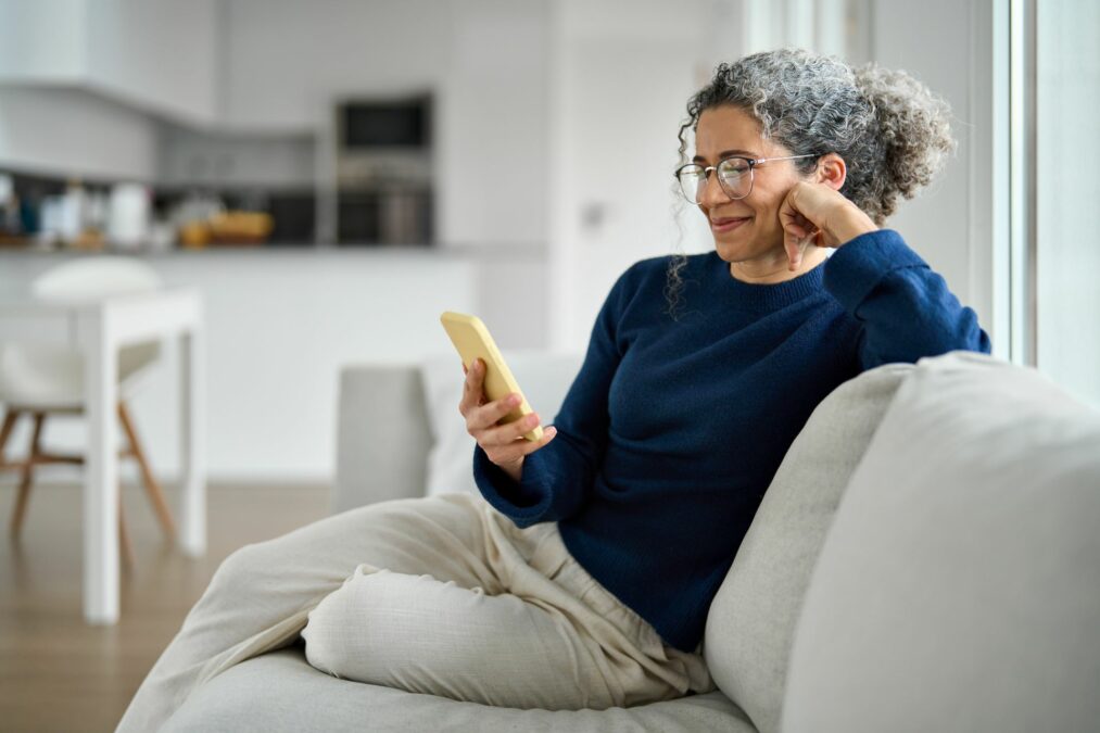 Happy middle aged older woman using smartphone sitting on couch at home.